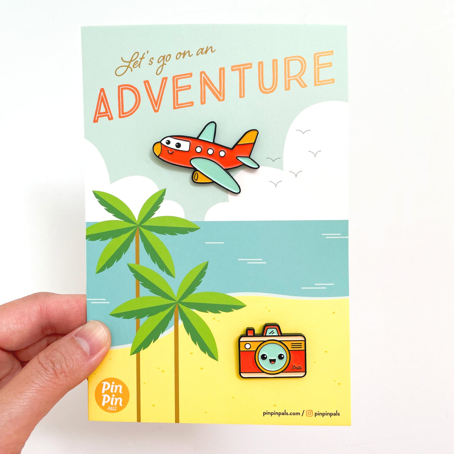 Hand holding a Let's Go on an Adventure postcard with plane and camera enamel pins